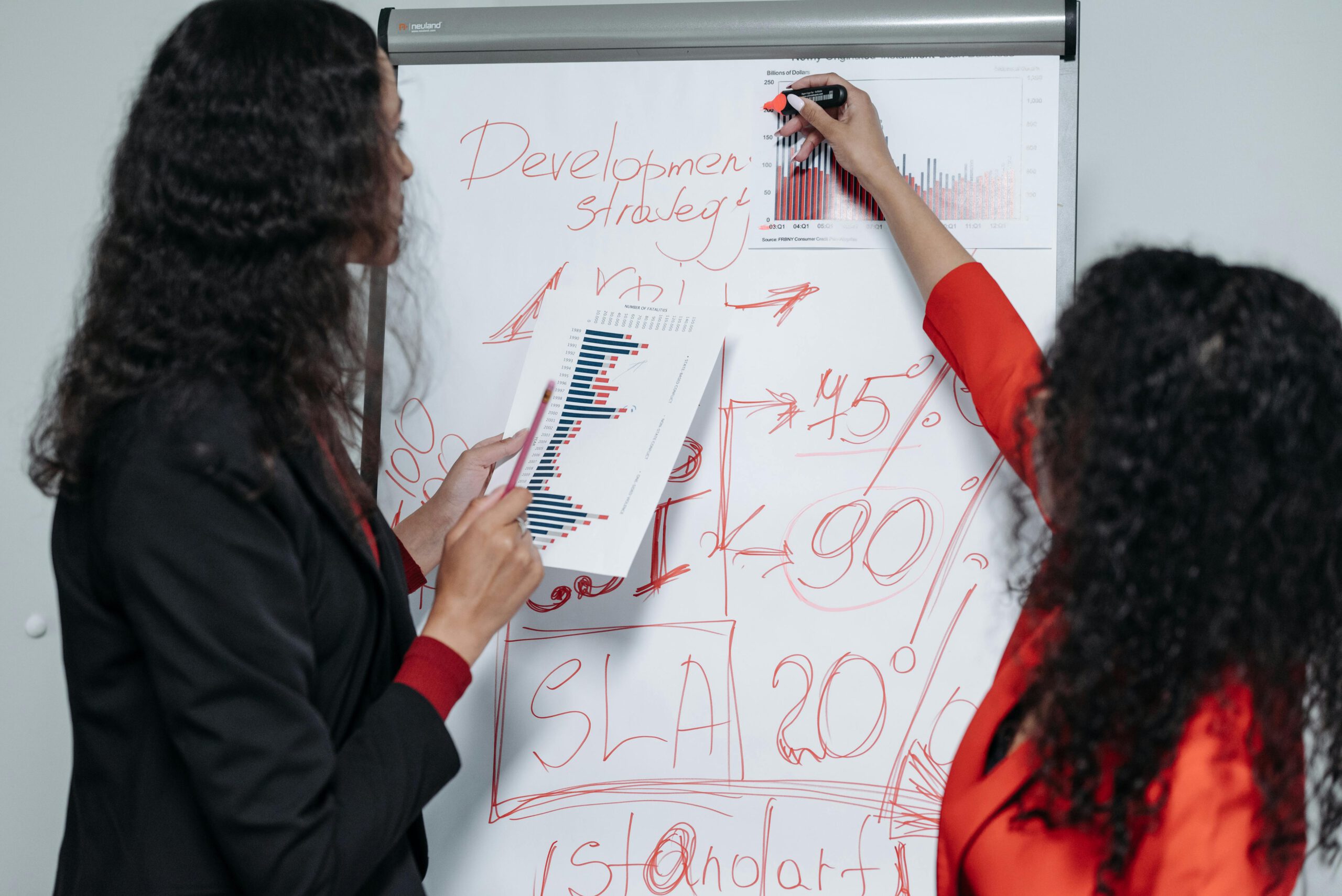 Two professional women discuss and strategize on business development in a modern office setting.